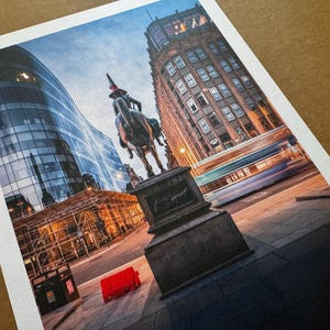 May include: A print depicting a city street with a statue of a person on horseback. Modern glass buildings and older architecture are visible. A red object is on the pavement. A blurred bus passes by.