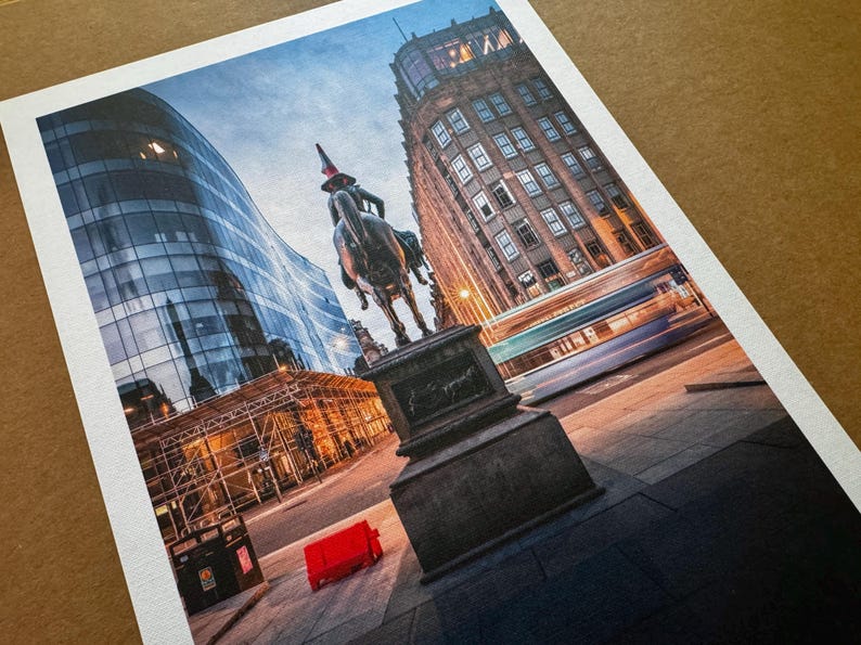 May include: A print depicting a city street with a statue of a person on horseback. Modern glass buildings and older architecture are visible. A red object is on the pavement. A blurred bus passes by.
