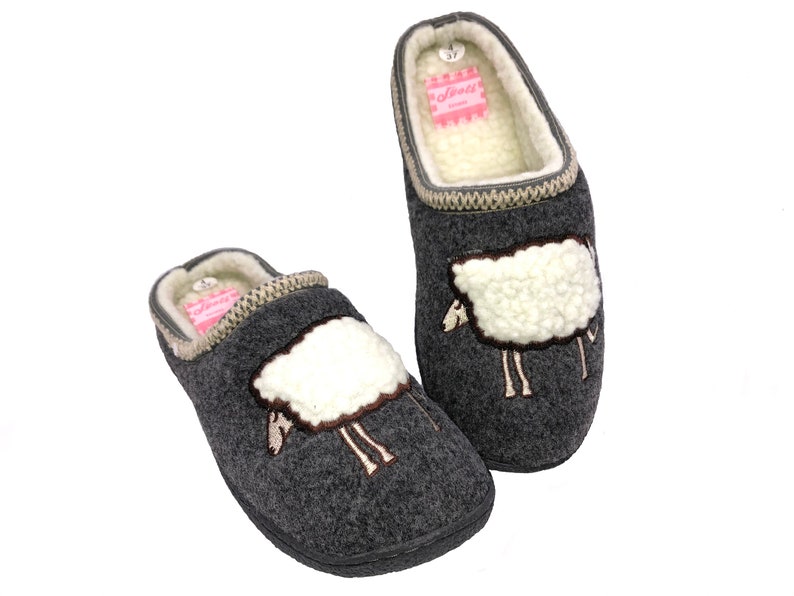 May include: A pair of gray wool slippers with a sheep design. The slippers have a white sheep with brown eyes and legs embroidered on the front. The slippers have a beige trim around the top and a pink label on the inside.