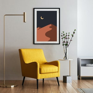 May include: A yellow armchair with black legs sits in a living room with a black framed print of a desert landscape with a crescent moon, a small white side table with a vase of pink flowers, and a white entertainment center with a black TV.