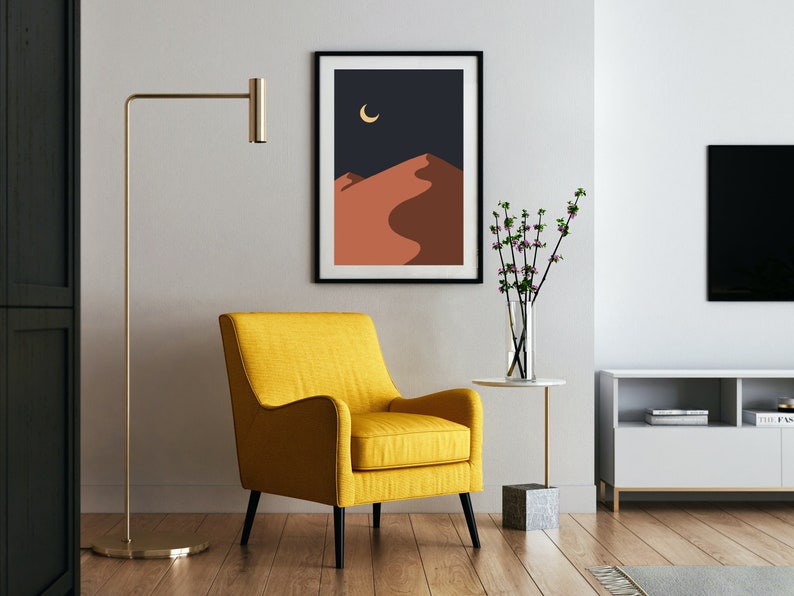 May include: A yellow armchair with black legs sits in a living room with a black framed print of a desert landscape with a crescent moon, a small white side table with a vase of pink flowers, and a white entertainment center with a black TV.