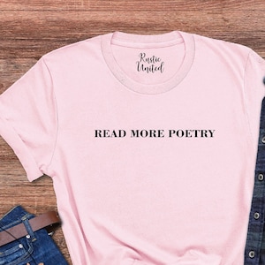 Op de afbeelding: Een roze T-shirt met de tekst "READ MORE POETRY" in zwarte letters. Het shirt ligt op een houten oppervlak met een blauwe spijkerbroek, een bruine leren riem, een zonnebril en een geruit overhemd.