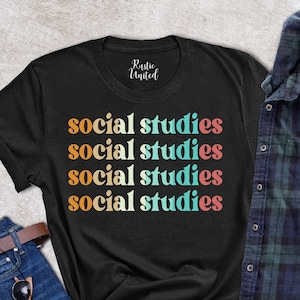 May include: Black t-shirt with the words "social studies" in a stacked design with retro-style lettering in orange, teal, white, pink, and orange. The shirt is paired with blue jeans, sunglasses, a brown wallet, a succulent, and a plaid shirt.