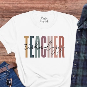 Pu&ograve; includere: Maglietta bianca con il testo "TEACHER technology" in strisce verticali colorate. La maglietta &egrave; stesa su una superficie di legno con un paio di jeans, occhiali da sole e una camicia a quadri.