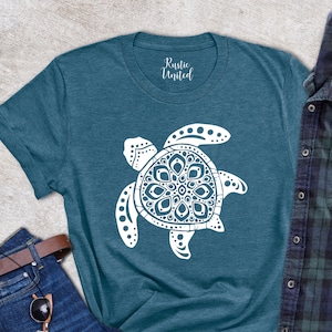 May include: Teal t-shirt with a white turtle design, paired with blue jeans, a brown belt, sunglasses, and a wallet. A blue and green plaid shirt is folded next to the t-shirt. A small succulent plant is also visible.
