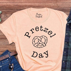 Op de afbeelding: Een perzikkleurig T-shirt met de tekst "Pretzel Day" en een zwart-witte illustratie van een pretzel.