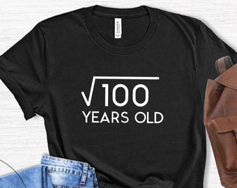 May include: Black t-shirt with the text "√100 YEARS OLD" in white. The shirt is paired with blue jeans, brown leather shoes, and a brown leather backpack. The items are arranged on a light wood-grain surface.