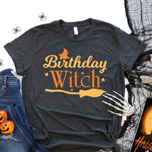 May include: A dark grey t-shirt with the text "Birthday Witch" in orange and yellow lettering. The shirt features a witch's hat and broom graphic. The shirt is displayed on a white background with jeans, a skull, and a Halloween party sign.