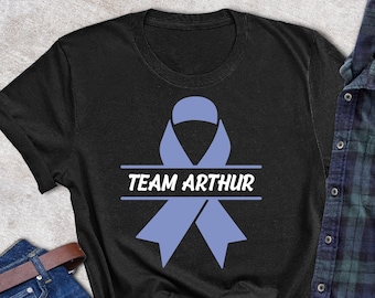 Op de afbeelding: Zwart T-shirt met een blauw lint en de tekst "TEAM ARTHUR" erop gedrukt. Het shirt ligt op een grijze ondergrond met een spijkerbroek, een zonnebril en een geruit overhemd.