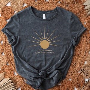 May include: A dark gray t-shirt with a gold sun graphic and the text "IV-VIII-MMXXIV SOLAR ECLIPSE TOTALITY".