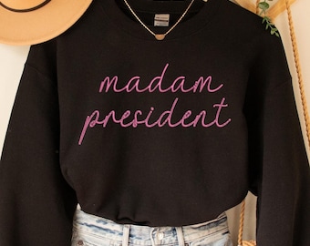 May include: A black sweatshirt with the words "madam president" in pink script is displayed with denim shorts and a tan hat. A small necklace is visible at the neckline. A woven bag hangs to the right.