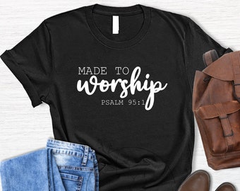 May include: Black t-shirt with white text that reads "MADE TO worship PSALM 95:1". The shirt is laid flat on a white wooden surface with a pair of blue jeans, a brown leather backpack, and brown leather boots.