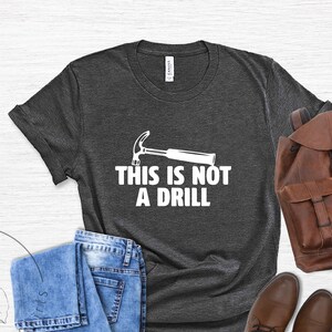 May include: A dark gray t-shirt with a white hammer and the text "This is not a drill" printed on the front. The shirt is displayed on a white wooden background with a pair of blue jeans, a brown leather backpack, and brown leather boots.