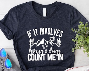 May include: Dark navy blue t-shirt with the text "IF IT INVOLVES hiking & dogs COUNT ME IN" in white. The graphic includes mountains, trees, a sun, and paw prints. The shirt is paired with gray jeans, blue sunglasses, and white sneakers.