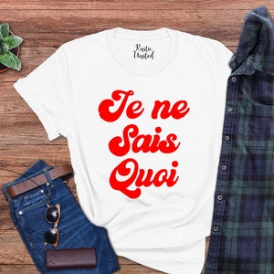 May include: White t-shirt with red text that says "Je ne sais quoi" in a retro font style. The shirt is laid out on a wooden surface with a pair of jeans and a plaid shirt.