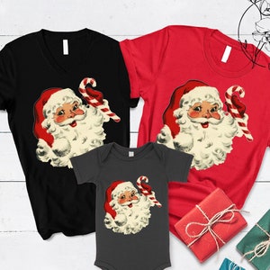 May include: A set of three Christmas shirts featuring a vintage illustration of Santa Claus holding a candy cane. The shirts are black, red, and gray.