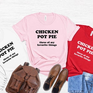 May include: Three t-shirts in white, pink, and red, each with the text "CHICKEN POT PIE three of my favorite things." A brown leather backpack, brown shoes, and blue jeans are also in the image. The shirts are from Orchid Shirts.