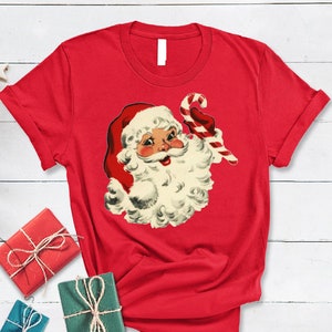 May include: Red t-shirt with a vintage illustration of Santa Claus holding a candy cane. The illustration is in red, white, and black.