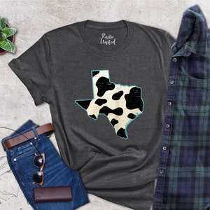 May include: A dark gray t-shirt with a white and black cow print outline of the state of Texas. The shirt is laid flat on a light gray textured surface with a pair of blue jeans, a brown leather belt, sunglasses, and a brown leather wallet.