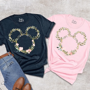 Peut inclure: Deux t-shirts, un bleu marine et un rose, chacun avec un motif floral de t&ecirc;te de Mickey Mouse. Le motif est compos&eacute; de feuilles vertes, d'accents dor&eacute;s et de fleurs roses. Les chemises sont pr&eacute;sent&eacute;es avec un jean et des chaussures marron.