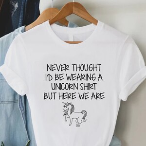 May include: White t-shirt with a black graphic of a unicorn and the text "Never thought I'd be wearing a unicorn shirt but here we are."