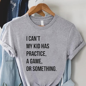 May include: A heather gray t-shirt with the text "I can't, my kid has practice, a game, or something." The shirt is hanging on a wooden hanger. A denim shirt and a hat are visible in the background.