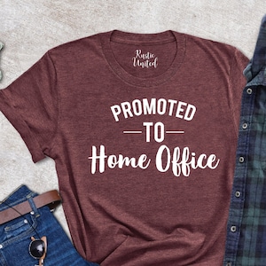 May include: A maroon t-shirt with the text "PROMOTED - TO - Home Office" in white, alongside blue jeans, a brown belt, sunglasses, and a brown wallet. A green and navy plaid shirt is also present. A small potted succulent is visible.