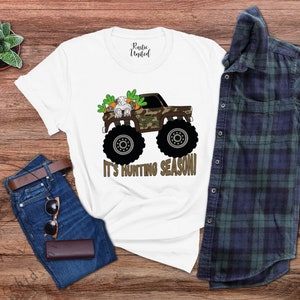 May include: White t-shirt featuring a monster truck with a camouflage design, a bunny, and the text "IT'S HUNTING SEASON!". The shirt is styled with blue jeans, a brown belt, sunglasses, and a wallet. A blue and green checked shirt is also present.