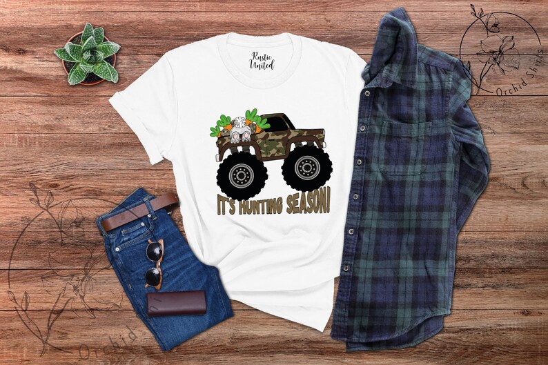 May include: White t-shirt featuring a monster truck with a camouflage design, a bunny, and the text "IT'S HUNTING SEASON!". The shirt is styled with blue jeans, a brown belt, sunglasses, and a wallet. A blue and green checked shirt is also present.