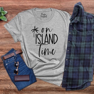 May include: A gray t-shirt with the text "*on island time" printed in black. The shirt is laid out on a wooden surface with a pair of jeans, sunglasses, and a brown leather wallet.