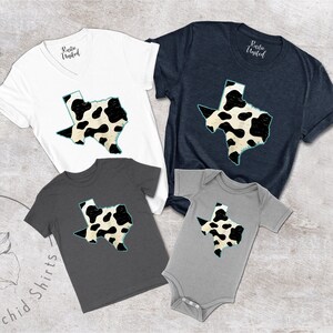 May include: A set of four shirts featuring a black and white cow print design of the state of Texas. The shirts are in various sizes and colors, including white, navy blue, and gray.