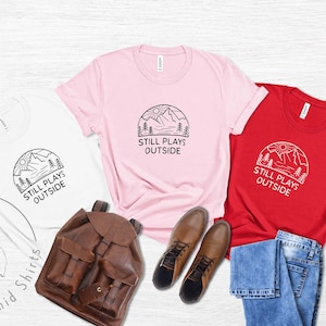 May include: Three t-shirts with a mountain scene and the text "Still Plays Outside". The shirts are white, pink, and red. The shirts are laid out on a white wooden surface with a brown leather backpack, blue jeans, and brown boots.