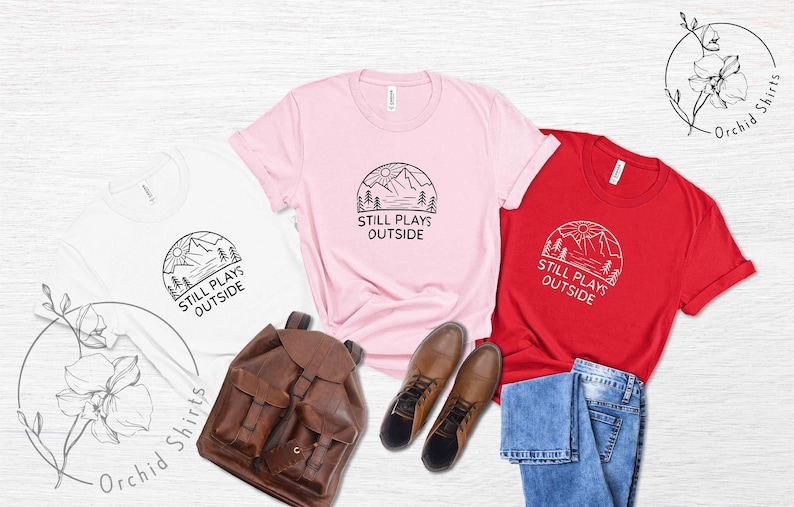May include: Three t-shirts with a mountain scene and the text "Still Plays Outside". The shirts are white, pink, and red. The shirts are laid out on a white wooden surface with a brown leather backpack, blue jeans, and brown boots.