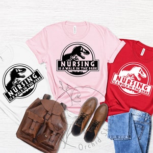May include: Three t-shirts with a dinosaur graphic and the text "Nursing is a walk in the park". The shirts are white, pink, and red.