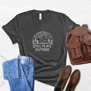 May include: A dark gray t-shirt with a white graphic of a mountain range, sun, and trees. The text "Still Plays Outside" is printed below the graphic.