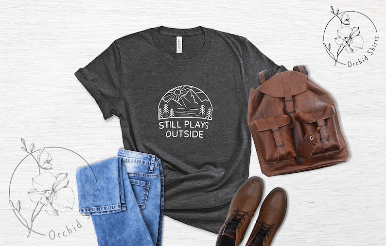 May include: A dark gray t-shirt with a white graphic of a mountain range, sun, and trees. The text "Still Plays Outside" is printed below the graphic.