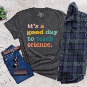 Op de afbeelding: Een donkergrijs T-shirt met de tekst "it's a good day to teach science." in kleurrijke letters. De tekst is in een retro-stijl lettertype.