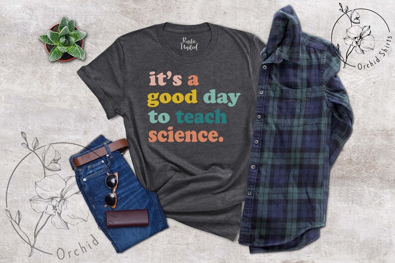 Op de afbeelding: Een donkergrijs T-shirt met de tekst "it's a good day to teach science." in kleurrijke letters. De tekst is in een retro-stijl lettertype.
