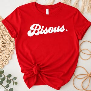 May include: A bright red short-sleeved t-shirt featuring the word Bisous. printed in a bold white retro-style script font across the center of the chest. The garment has a crew neckline and is tied in a knot at the bottom front hem.