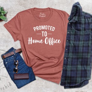 May include: A terracotta-coloured t-shirt with the text "PROMOTED TO - Home Office" in white, paired with blue jeans, a brown belt, sunglasses, and a wallet. A green and blue plaid shirt is also included.