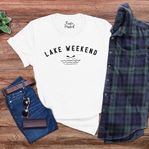 以下が含まれることがあります： 「Lake Weekend」の黒い文字が入った白いTシャツ。湖とオールのグラフィックが特徴です。シャツは、青いジーンズ、茶色のベルト、サングラス、財布、チェック柄のシャツと組み合わせています。小さな多肉植物も見えます。