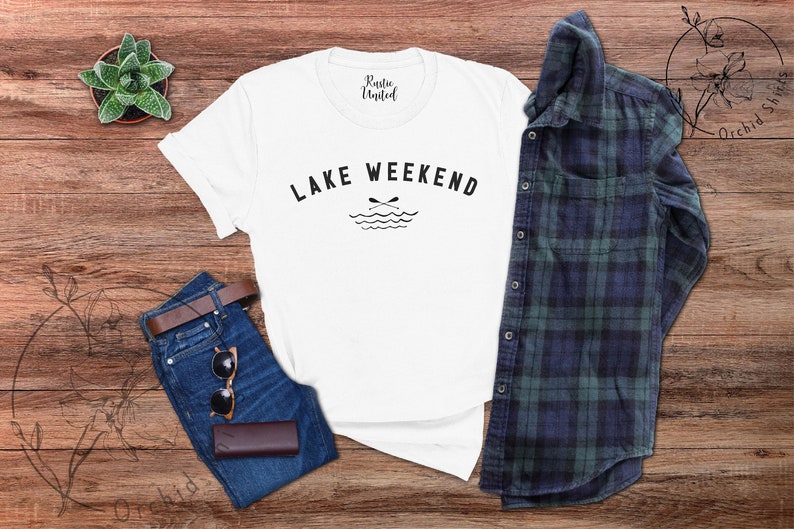 以下が含まれることがあります： 「Lake Weekend」の黒い文字が入った白いTシャツ。湖とオールのグラフィックが特徴です。シャツは、青いジーンズ、茶色のベルト、サングラス、財布、チェック柄のシャツと組み合わせています。小さな多肉植物も見えます。