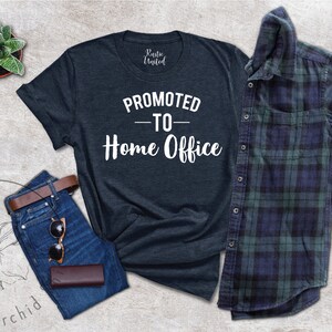 May include: A dark navy blue t-shirt with the white text "PROMOTED - TO - Home Office". The image includes blue jeans, a brown belt, sunglasses, and a brown wallet. A green and blue checked shirt is also present.