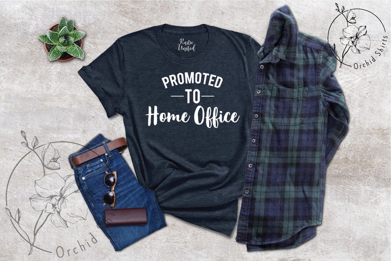 May include: A dark navy blue t-shirt with the white text "PROMOTED - TO - Home Office". The image includes blue jeans, a brown belt, sunglasses, and a brown wallet. A green and blue checked shirt is also present.