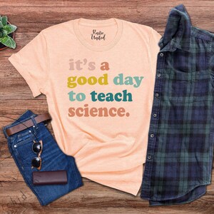 Op de afbeelding: Een perzikkleurig T-shirt met de tekst "it's a good day to teach science." in kleurrijke letters. Het shirt ligt op een houten oppervlak met een spijkerbroek, een zonnebril en een geruit overhemd.