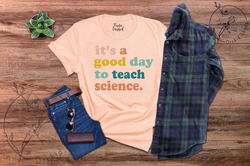 Op de afbeelding: Een perzikkleurig T-shirt met de tekst "it's a good day to teach science." in kleurrijke letters. Het shirt ligt op een houten oppervlak met een spijkerbroek, een zonnebril en een geruit overhemd.