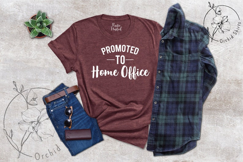 May include: A maroon t-shirt with the text "PROMOTED - TO - Home Office" in white, alongside blue jeans, a brown belt, sunglasses, and a brown wallet. A green and navy plaid shirt is also present. A small potted succulent is visible.