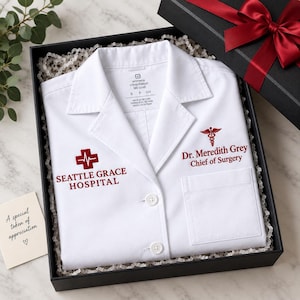 May include: A white lab coat folded in a black gift box with a red ribbon. The coat features red embroidery reading Seattle Grace Hospital with a medical cross, and Dr. Meredith Grey Chief of Surgery with a caduceus symbol on the chest pocket.