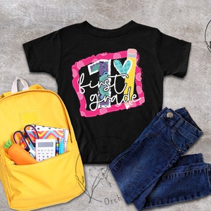 May include: A black t-shirt with a pink and white graphic that says "1st grade" with a pencil and a heart. A yellow backpack with school supplies is in the background.