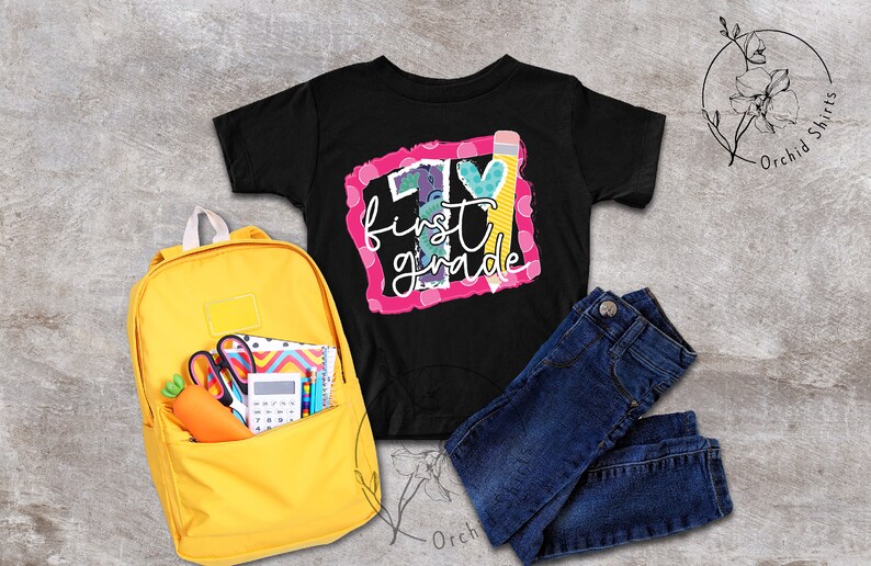 May include: A black t-shirt with a pink and white graphic that says "1st grade" with a pencil and a heart. A yellow backpack with school supplies is in the background.
